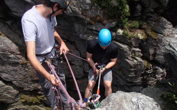 Two people wearing safety gear are secured by ropes as they prepare to rappel down a cliff. One person appears to be an instructor, giving direction to the other person.